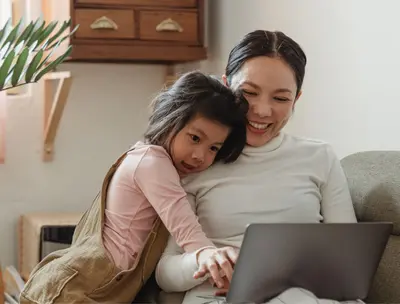Mother working on a laptop at home while her young daughter leans in close beside her