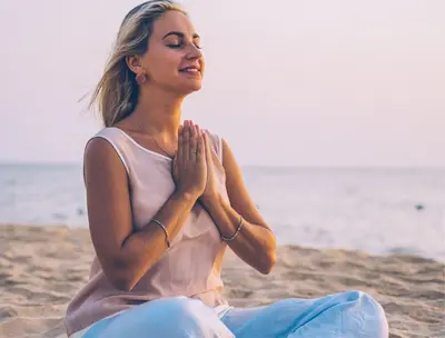 Woman meditating cross-legged on a beach, hands in prayer and eyes closed, with the ocean behind her