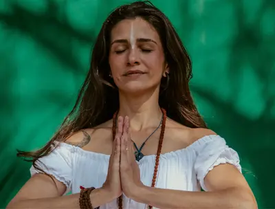 Woman with hands pressed together in prayer, eyes closed, against greenery