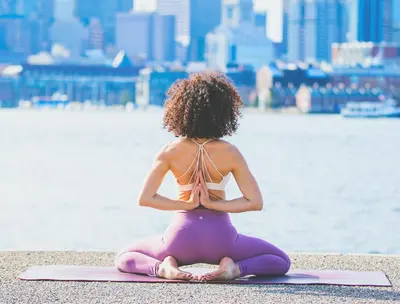 Woman sitting in a yoga pose on a waterfront promenade with the city skyline behind her