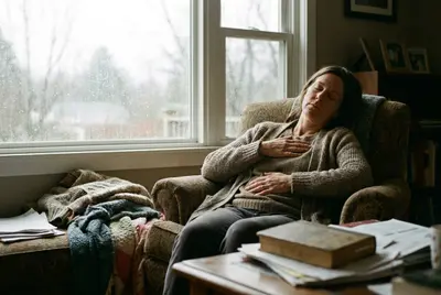 Woman in an armchair by a rainy window, eyes closed, one hand on her chest, breathing slowly