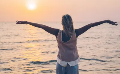 Woman standing with arms outstretched facing the ocean at sunset