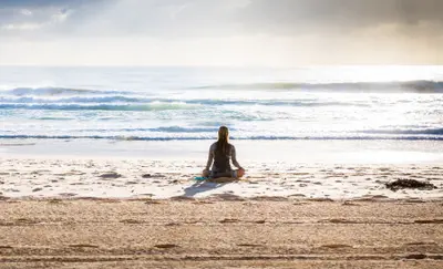 Woman sitting cross-legged on the beach, meditating and facing the ocean