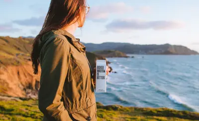 Woman standing on a coastal cliff holding a carton of boxed water, looking out at the ocean