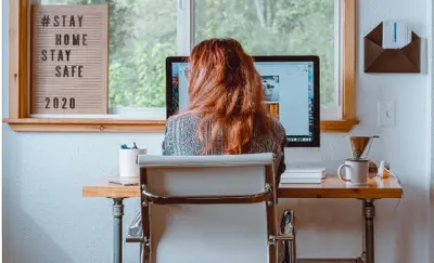 Woman working at a home desk facing her computer, with a "Stay Home Stay Safe 2020" sign on the windowsill