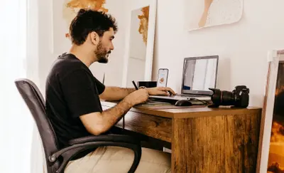 Man working at a wooden desk with a laptop, camera, and framed art on the wall